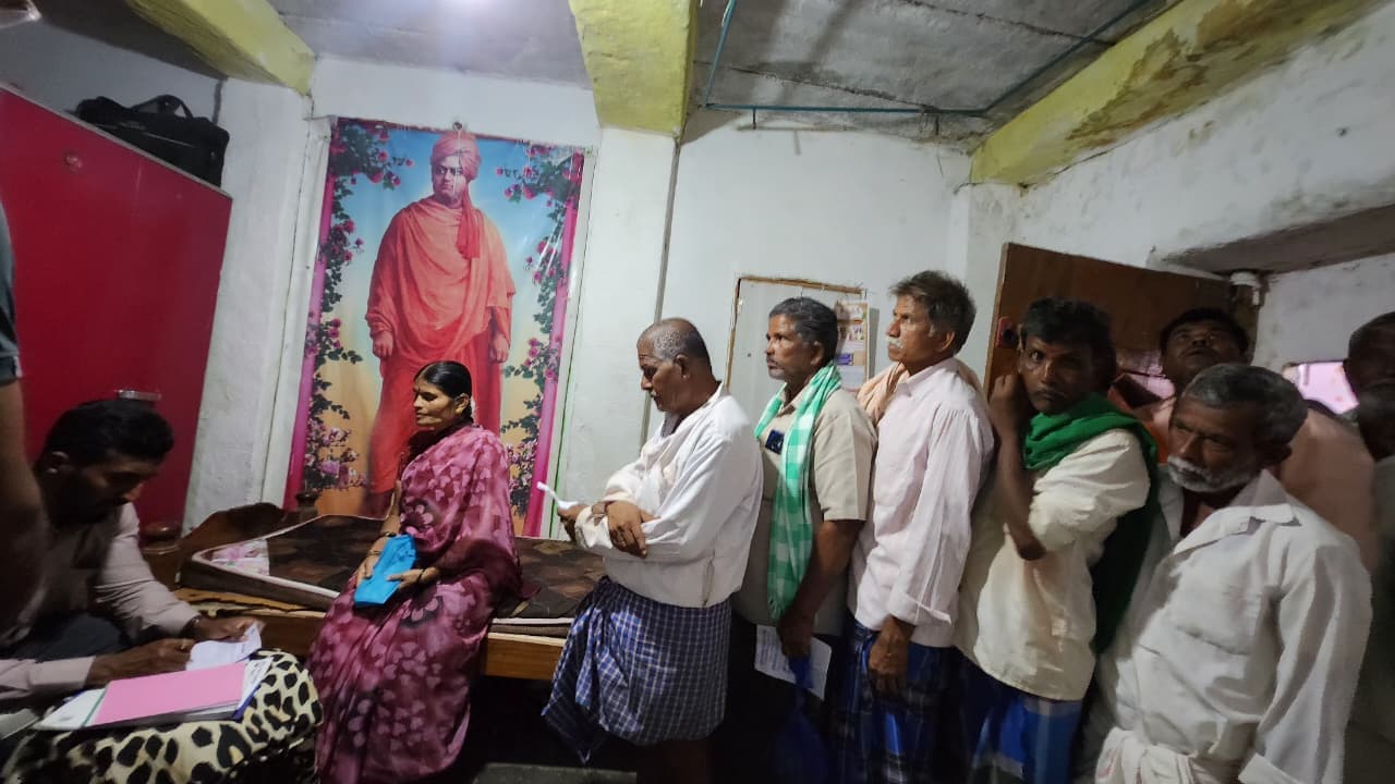 Rural villagers queuing for doctor consultation in village clinic hall under Swami Vivekananda portrait